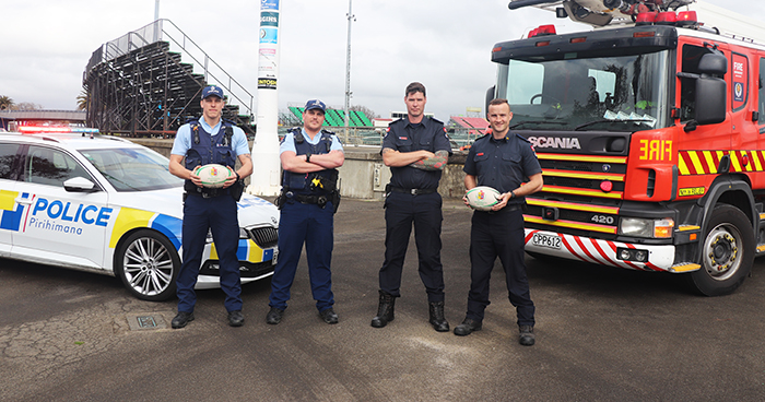 Police and FENZ staff holding rugby balls in front of a police car and fire truck.