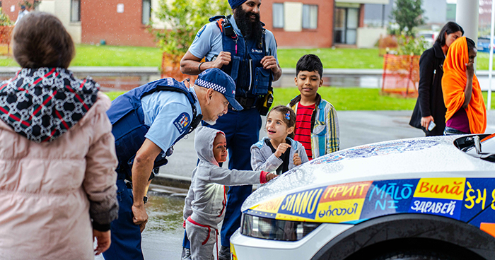 A police officer leaning forward and listening to some young children who are stood next to Police's ethnic car and pointing at some of the greetings in different languages that cover the car.