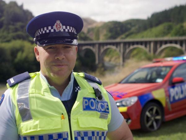 Senior Constable Tony Sharp standing in front of a red police car. Photo: Highway Cops/TVNZ