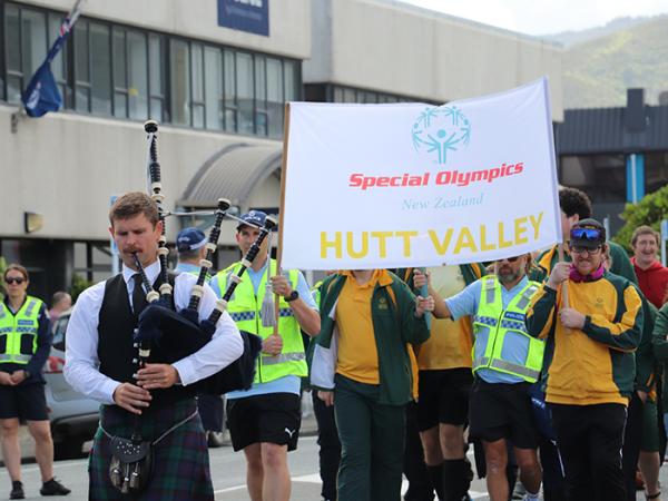 A bagpiper leads a police officer and athlete who are holding the Hutt Valley banner high followed by athletes.