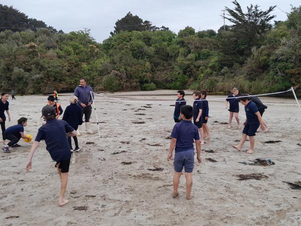 A group of about ten kids playing volleyball while adults watch on on either side of the net.