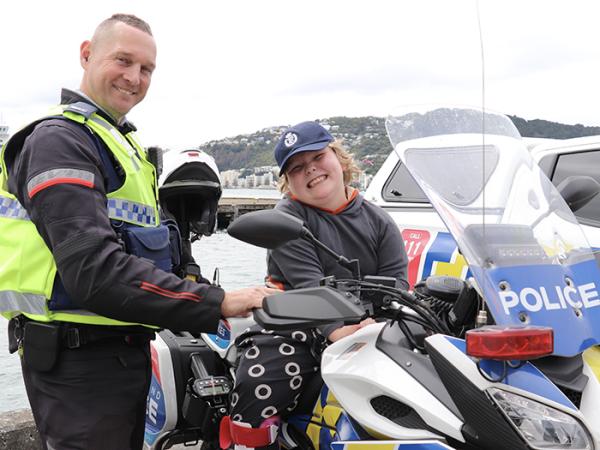 Elliot sitting on the police motorcycle, looking at the camera and smiling next to Constable Ash Williams.