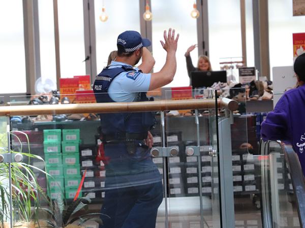 Photo taken from behind two police officers waving back to a retailer in a shop who's waving hello to them in a shopping mall. 