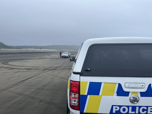 Close up of part of the back of a police vehicle, with officers talking to people in a car in the distance, on the beach.