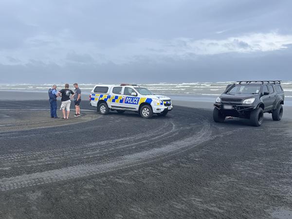 Police talking to the two people on the beach with a police car and a black truck parked close by on the sand.