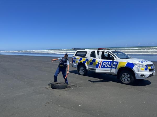 A police officer approaching a discarded tyre on the beach, with their police vehicle in the background.