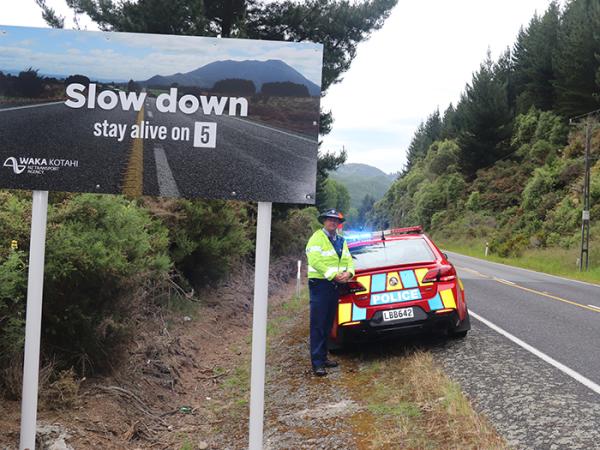 Steven standing behind a police car next to one of the 'Stay Alive on 5' signs on the busy Napier-Taupo Road.
